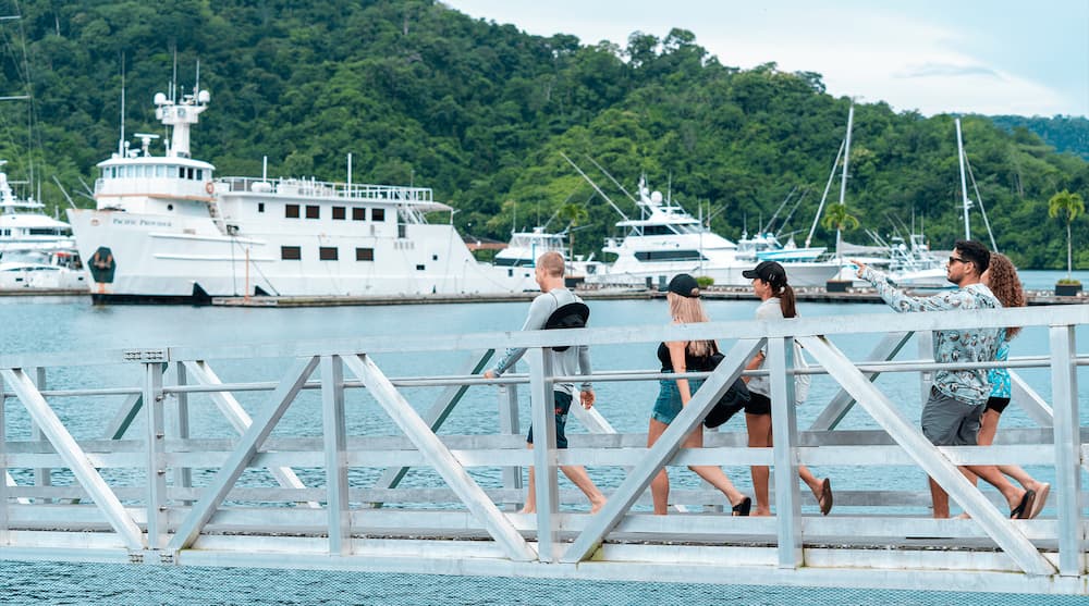 Visitors walking a dock at Marina Bahia Golfito with sportfishing yachts and rainforest-covered mountains behind them in Golfito, Costa Rica.
